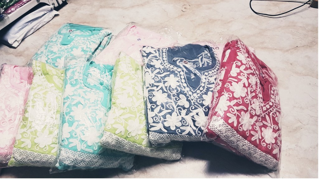 A row of multi-coloured kurtas in the same design. They are folded and wrapped inside transparent plastic packaging. They are lying on a tiled beige floor. Each has heavy white embroidery around the neckline. Each is resting on top of the previous one, sliding off towards the floor. Starting from the left, there is pale pink, lime green, turquoise blue, lime green, navy blue, and bright red one.